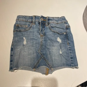 Blå jeanskjol från 157 Neville - Snygg blå jeanskjol från 157 Neville i klassisk denim med slitna detaljer och rå kant nertill. Kjolen har fem fickor, bälteshällor och knappstängning framtill. Perfekt för dig som gillar en avslappnad och trendig stil.