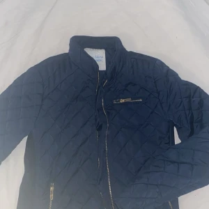 Mörkblå höstjacka - Quiltad mörkblå jacka från Samosa med dragkedjor i silver och ståkrage. Jackan har ett rutigt mönster, lång ärm och flera fickor med dragkedja. Perfekt för dig som gillar en stilren stockholmslook.