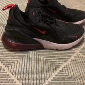 Nike Air Max 270 svarta sneakers - Svarta Nike Air Max 270 sneakers med röda detaljer och logga. Skorna har en luftig mesh-ovandel, snörning och en tydlig Air Max-sula i rött och svart. Vit mellansula och röd hälflik ger en sportig look. Perfekta för dig som gillar streetwear och snygga detaljer.
