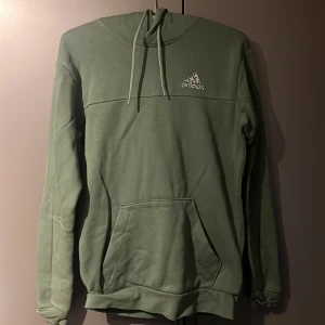 Grön hoodie från Adidas - Säljer en grön hoodie från Adidas med klassisk logga på bröstet. Tröjan har huva med snörning och en stor magficka framtill. Perfekt för chill dagar eller när du vill ha en avslappnad stil.