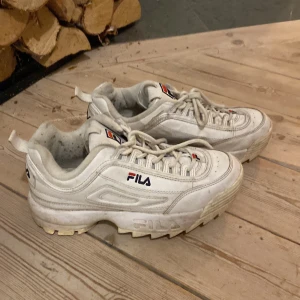 Vita chunky sneakers från Fila - Säljer ett par klassiska vita Fila sneakers med chunky sula och snörning. Skorna har Fila-logga på sidan, tungan och hälen. Tillverkade i syntetmaterial och skinnimitation, med rund tå och platt sula. Perfekta för dig som gillar retrostil och streetwear. Storlek 38, hör av dig om du har frågor eller funderingar!