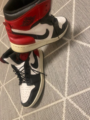 Nike Air Jordan 1 High röd/svart/vit - Nike Air Jordan 1 High sneakers i klassisk färgkombination med rött, svart och vitt. Skorna har högt skaft, snörning och perforerad tå. Ikonisk Jumpman-logga på plösen och Air Jordan Wings på sidan. Tillverkade i skinn med platt sula och rund tå.
