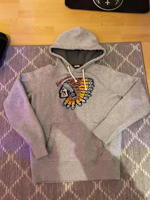 Grå hoodie Abercrombie & Fitch - Snygg grå hoodie från Abercrombie & Fitch med broderad färgglad indianfjäder på bröstet och texten 'Abercrombie'. Mjuk insida, stor huva med vita snören och ribbade muddar. Klassisk känguruficka framtill. Perfekt för chill och streetwear. (Den ser ut å vara rätt så använd men den designen är att den ska vara lite sliten å så🤟🏽🤟🏽)