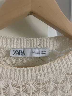 Vit spetsblus med volang från Zara - Supersöt vit blus från Zara med korta ärmar, spetsdetaljer och volangkant nertill. Blusen har ett vackert virkat mönster och rund halsringning. Perfekt för dig som gillar romantisk och feminin stil.
