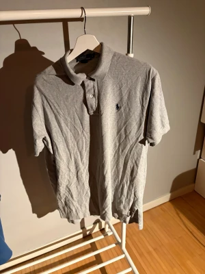 Grå pikétröja från Polo Ralph Lauren - Klassisk grå pikétröja från Polo Ralph Lauren med broderad marinblå logga på bröstet. Tröjan har korta ärmar, krage och knappar framtill. Tillverkad i mjuk bomull som känns skön mot huden.