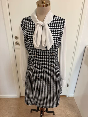 Svartvit klänning med knytblus från SHEIN - Stilren svartvit klänning från SHEIN med klassiskt hundtandsmönster och vita långärmade ärmar i lätt material. Framtill finns en snygg knytblusdetalj vid halsen och dekorativa knappar. Perfekt för dig som gillar en chic och tidlös look.