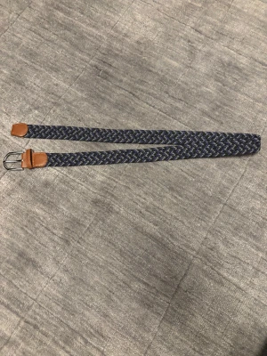 Mörkblå flätad bälte med bruna detaljer - Stilrent flätat bälte, i blå och grå färg, fina detaljer och mönster, perfekt till alla jeans, Grisch.          Hör av dig vid minsta intresse!