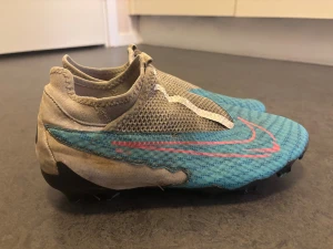 Nike Phantom GT2 blå fotbollsskor - Nike Phantom GT2 fotbollsskor i blått med mönstrad ovandel och rosa swoosh. Skorna har en strömlinjeformad design utan snörning, meshpaneler för ventilation och svarta dobbar på sulan för bra grepp på planen. Perfekt för dig som vill sticka ut med färg och stil på matchen.