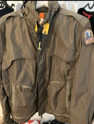 Parajumpers mörkgrön vindjacka M - Säljer en mörkgrön vindjacka från Parajumpers i storlek M. Jackan har huva, coola patchar på ärmen, dragkedja framtill och flera fickor med zip. Materialet är lätt och syntetiskt, perfekt för blåsiga dagar. Snygga detaljer med gul rem och meshfoder.