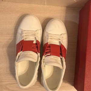 Valentino opens röda - Säljer ett par vita sneakers från Valentino Garavani med bred röd läderdetalj över snörningen. Skorna har rund tå och platt sula, och levereras med originalkartong och dustbag. Perfekta för dig som gillar exklusiva och stilrena sneakers. Lite smutsiga ifram men det kan jag ta bort innan jag skickat de