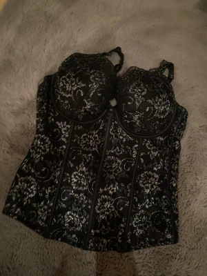 Svart glittrig korsett med blommönster - Säljer en svart korsett med silverglittrigt blommönster och markerade sömmar. Toppen har formade kupor och justerbara axelband. Materialet är glittrigt och ger en lyxig vibe, perfekt för att sticka ut. Blommönstret är detaljerat och hela korsetten har en snygg, tight passform.