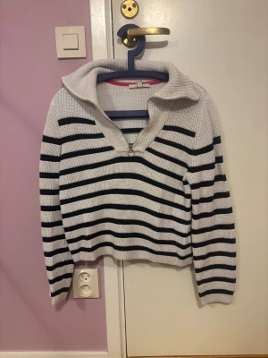 Randig stickad tröja med krage - Säljer en vit och svart stickad tröja med bred krage och half zip framtill. Tröjan har ett snyggt randigt mönster och långa ärmar, perfekt för dig som gillar chill och stilren look. Passar dig som vill ha en mysig och trendig tröja till garderoben.