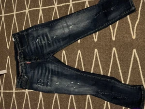 Mörkblå Dsquared2 jeans med slitningar - Snygga mörkblå jeans från Dsquared2 med coola slitningar och färgstänk på framsidan. Klassisk femficksmodell med Dsquared2-logga på bakfickan. Jeansen har en smal passform och är tillverkade i jeansmaterial med lätt stretch.