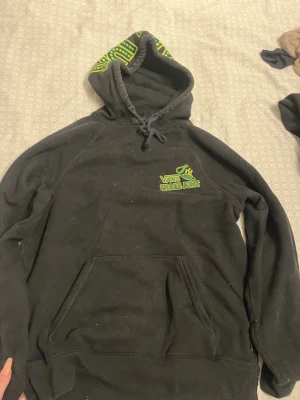 Svart Vans Shake Junt hoodie S - Svart hoodie från Vans x Shake Junt i storlek small. Framsidan har broderad logga i grönt och gult, baksidan har stort tryck med texten 'Getchu some chicken & waffles' i samma färger. Huva med snörning och tryck på insidan, samt klassisk magficka. Mjuk bomullsmix.