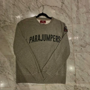 Grå sweatshirt från Parajumpers - Säljer en grå sweatshirt från Parajumpers med stort svart tryck över bröstet och patch på ena ärmen. Tröjan har rund hals, långa ärmar och ribbade muddar vid ärmslut och nederkant. Perfekt för chill dagar och enkel att matcha. (SKICK-bra)