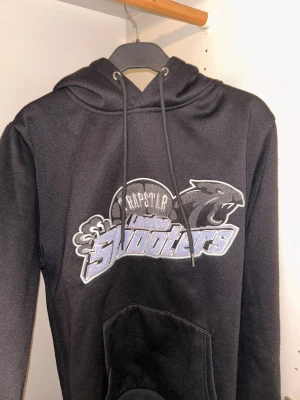 Svart Trapstar hoodie med tryck - Säljer en svart hoodie från Trapstar med stort broderat och tryckt motiv på bröstet. Tröjan har huva med snörning och en klassisk känguruficka framtill. Perfekt för dig som gillar streetwear och vill sticka ut med cool grafik.