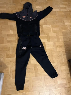 Nike tech x syna world tracksuit  - Nike tracksuit  i svart med rosa logga och grafiskt tryck på bröstet. Tröjan har huva, hel dragkedja och två fickor med dragkedja. Materialet är mjukt och känns som bomull och polyester. Snygg kontrast med grått upptill och rosa detaljer. Perfekt för chill eller träning.