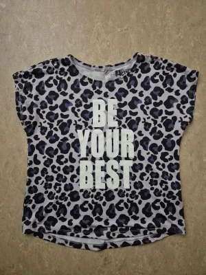 T-shirt - Cool t-shirt med leopardmönster i svart och mörklila på ljusgrå botten. Stor vit text 'BE YOUR BEST' framtill. Kortärmad och tillverkad i mjuk polyester, perfekt för dig som gillar att sticka ut. Rund hals och rak passform.