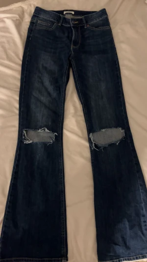 Ihåliga Jeansbyxor -mörkblåa  - Säljer dessa otroligt fina mörkblå jeans med hål i knäna och utsvängd modell (bootcut). Superbekväma, passar till nästan allt i garderoben och i riktigt bra skick. Inga slitage eller fläckar. Storlek 36. 
