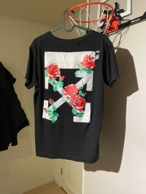 Svart 0ff White t-shirt med rosor - Svart t-shirt från 0ff-White med stort vitt logotryck och röda rosor på ryggen. Framtill finns små detaljer med samma ros- och logomotiv vid nederkanten. T-shirten har rund hals och korta ärmar, tillverkad i mjuk bomull för skön passform.