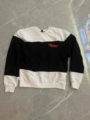 Svartvit sweatshirt från Mavi - Loose fit sweatshirt från Mavi i svart och vitt med breda blockränder och röd textdetalj på bröstet. Tröjan har rund halsringning, ribbade muddar och är tillverkad i mjukt material som känns skönt mot huden. Perfekt för en chill och stilren lock.