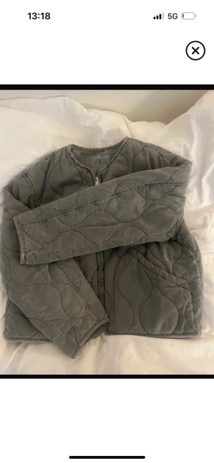 Grå quiltad jacka från Zara - Quiltad jacka från Zara i grått med vågigt mönster. Jackan har rund hals, lång ärm och dragkedja framtill. Den har en loose passform och är perfekt för lager-på-lager. Materialet är mjukt och känns skönt mot huden.