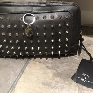 Svart väska med nitar från Friis & Company - Cool svart väska från Friis & Company, dekorerad med silverfärgade nitar. Väskan har en dragkedja upptill och en stilren design som passar perfekt för en edgy look.Har dock lite "skönhetsfel" som syns på bild.. Oanvänd med lappar kvar. 