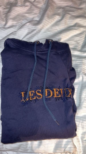 Mörkblå hoodie Les Deux med broderi - Säljer en mörkblå hoodie från Les Deux i storlek S. Tröjan har snyggt broderat Les Deux-tryck i gult och blått på bröstet, justerbar snörning vid huvan och en klassisk känguruficka framtill. Perfekt för dig som gillar stilrena streetwear-plagg.