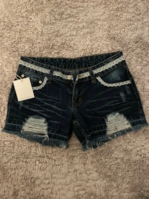 Mörkblå jeansshorts med spetsdetaljer - Snygga mörkblå jeansshorts med ljus spets längs midjan och fickorna. Shortsen har fransade kanter och slitna detaljer framtill för en coolt look. Klassisk femficksmodell med knapp och dragkedja. Prislapp finns även kvar!! Midjemått: 36cm. Skriv vid fler frågor❤️❤️🥰🥰