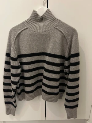 Grå och svart randig polotröja H&M - Säljer en grå stickad polotröja från H&M i storlek S. Tröjan har svarta horisontella ränder, hög ribbad krage och långa ärmar. Perfekt för dig som gillar en chill och klassisk look med lite edge.