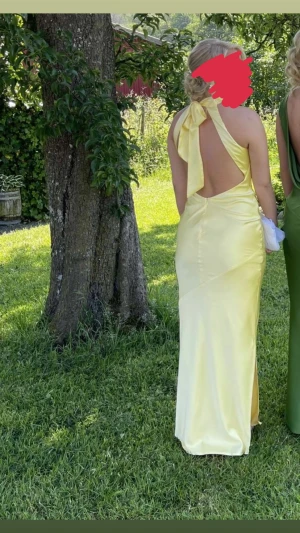 Gul långklänning med öppen rygg - Snygg gul långklänning i satin med halterneck och stor rosett i nacken. Klänningen har en öppen rygg och figurnära passform som framhäver siluetten. Bara använd en gång. Perfekt till bal eller bröllop🥰