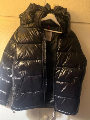 Moncler pufferjacka M - Svart pufferjacka från Moncler med glansig finish, huva och två sidofickor med dragkedja. Jackan har quiltad design och innerfoder med serietidningsmönster. Perfekt för kalla dagar och har snygga detaljer som snörning i nederkant. Avtagbar luva även. Ej Äkta!