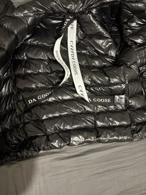 Svart dunjacka från Canada Goose - Säljer en svart, glansig dunjacka från Canada Goose med klassisk röd och vit logotyp på ärmen. Jackan har dragkedja, quiltad design och justerbara snören i nederkant. Perfekt för kalla dagar och har en riktigt snygg streetstil. Kvitto finns. Priset är ej hugget i sten. Nypris 10000 kr. Tagen finns kvar