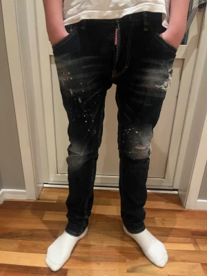 Dsquared2 mörkblå slim jeans - Snygga mörkblå jeans från Dsquared2 med slitningar, färgstänk och skinny passform. Jeansen har klassiska fem fickor, orange kontrastsömmar och en röd Dsquared2-tag vid gylfen. Perfekt för dig som gillar en edgy och trendig look. I storlek 44. Modelen är 173 cm lång. 