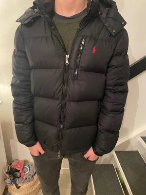 Svart dunjacka från Polo Ralph Lauren - Säljer en svart dunjacka från Polo Ralph Lauren med huva och dragkedja. Jackan har en klassisk pufferstil, röd broderad logga på bröstet och flera praktiska fickor. Perfekt för kalla dagar och riktigt eftertraktad just nu inom ungdomar