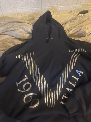 Mörkblå hoodie V1969 Italia - Snygg mörkblå hoodie från V1969 Italia med stort silvrigt tryck framtill. Tröjan har huva och ribbade muddar vid ärmslut och nederkant. Perfekt för dig som gillar streetstyle och vill sticka ut med en cool logga.