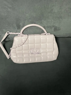 Vit quiltad handväska från V 1969 - Snygg vit handväska från V 1969 med quiltad design och stilren logga framtill. Väskan har både handtag och axelrem med kedjedetaljer, samt praktiskt innerfack med dragkedja. Perfekt storlek för dina essentials och ger en clean vibe till din outfit.