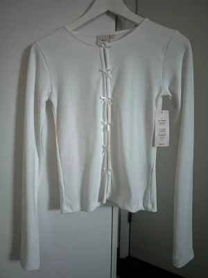 Vit ribbad topp med rosetter från Nelly - Supersöt vit ribbad topp från Nelly i storlek XS. Långärmad modell med flera små rosetter framtill som stängning. Tunn och stretchig bomullskänsla, perfekt för lager-på-lager eller som statement-piece. Enkel rund halsringning och figurnära passform. Ny med prislapp kvar