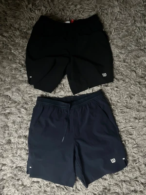Svarta och marinblå Wilson tränings shorts - Två par sportiga shorts från Wilson, ett par i svart och ett par i marinblått. Dem blåa har aldrig använts och dem svarta är endast använda ett par gånger. Båda är i ett mycket bra skick.