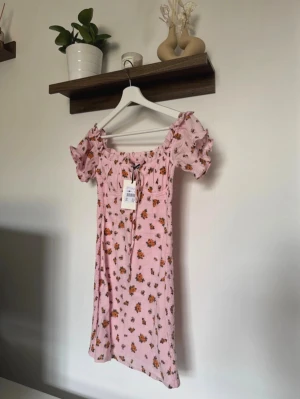 Rosa blommig klänning från Bik Bok - •Supersöt rosa kortklänning från Bik Bok med små orange och gröna blommor. •Klänningen har puffiga korta ärmar, smockad överdel och knyt framtill.  •Tillverkad i 100% bomull. Helt ny med prislappen kvar