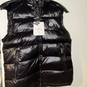 Svart dunväst från Moncler - Snygg svart dunväst från Moncler med glansig finish och huva. Västen har två praktiska fickor med dragkedja och Moncler-logga på bröstet. Perfekt för att addera lite lyx till din outfit och hålla dig varm under kyliga dagar.