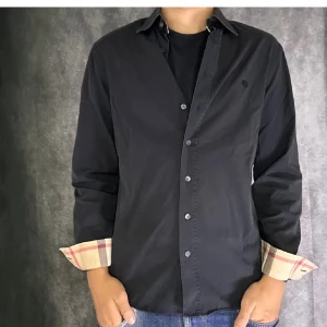 Svart Burberry Brit skjorta med rutig detalj - Svart skjorta från Burberry Brit med klassisk krage och knappar framtill. Insidan av manschetterna har den ikoniska Burberry-rutiga detaljen i beige, rött och grått. Skjortan är långärmad och har en normal passform, tillverkad i mjuk bomull. Perfekt för dig som gillar stilrena plagg med lyxig touch. M män passar även S, pris kan diskuteras!