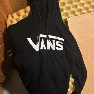 Svart hoodie från Vans med logga - Svart hoodie från Vans med stor vit logga framtill. Klassisk modell med huva och snörning, perfekt för en avslappnad streetstyle-look. Mjuk och bekväm, passar till jeans eller joggers.