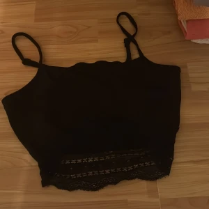 Svart spetsig linnetopp med smala band - Säljer en svart linnetopp med smala axelband och snygga spetsdetaljer längst ner. Toppen har en croppad passform och subtila ränder på insidan för extra stil. Perfekt för dig som gillar en enkel men ändå cool look.