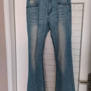 Ljusblå bootcut jeans från P.P.T Jeans - Säljer ett par ljusblå jeans från P.P.T Jeans med bootcut-modell. Jeansen har snygga slitningar och blekta partier framtill och baktill, klassiska fem fickor och normal passform. Perfekta för dig som gillar retrovibbar och vill ha ett par jeans med lite extra attityd.