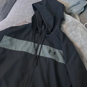 Svart vindjacka från Under Armour  - Svart vindjacka från Under Armour med grått band över bröstet 
