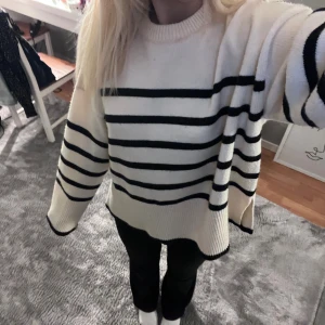 Randig stickad tröja i oversized modell - Säljer en vit stickad tröja med svarta horisontella ränder. Tröjan har en oversized passform, rund hals och långa ärmar med breda muddar. Perfekt för dig som gillar chill och avslappnad stil. Materialet är mjukt och skönt, och tröjan har slits i sidorna.