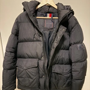 Svart pufferjacka från Tommy Hilfiger - Svart pufferjacka från Tommy Hilfiger med huva, stora fickor med lock och dragkedja framtill. Jackan har en clean look med diskret logga på ärmen och klassiska Hilfiger-detaljer i rött, vitt och blått på insidan. Perfekt för kalla dagar och riktigt snygg till streetwear.