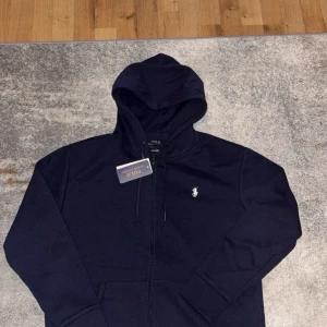 Marinblå hoodie från Polo Ralph Lauren - Snygg marinblå hoodie från Polo Ralph Lauren med klassisk vit logga på bröstet. Tröjan har huva med snörning, dragkedja framtill och två fickor. Perfekt för chill dagar och enkel att matcha med jeans eller joggers.