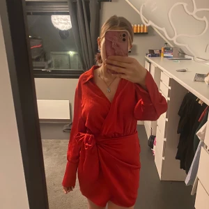 Röd omlottklänning med knyt - Säljer en röd kort omlottklänning med långa ärmar och snygg krage. Klänningen har ett brett knytband i midjan som ger en markerad siluett och en cool, avslappnad look. Perfekt för dig som gillar färg och vill sticka ut. Och speciellt nu till jul